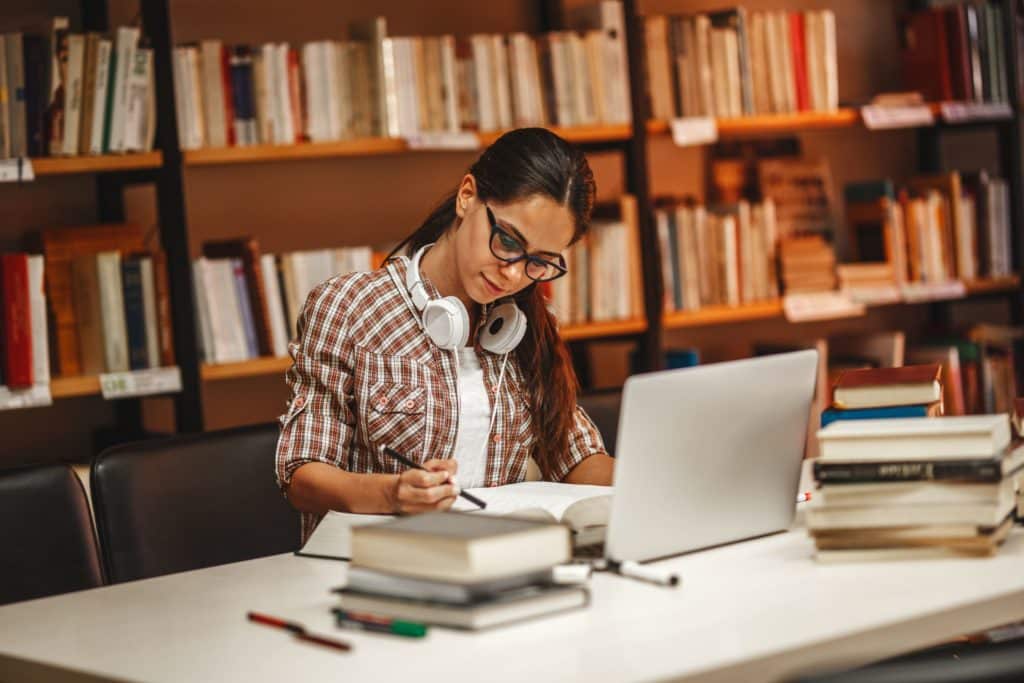 A woman sits in a library, studying with books and her laptop.