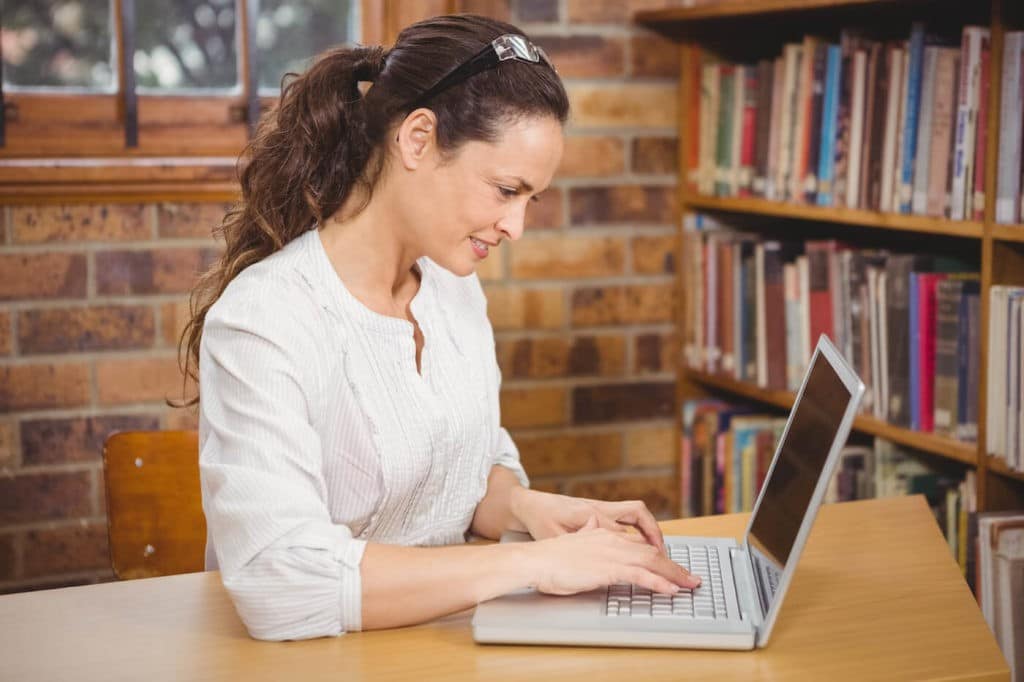 Teacher sitting at a table in the library working on a laptop.