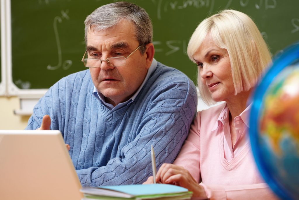 Two older teachers sitting in a classroom together reviewing data on a laptop.