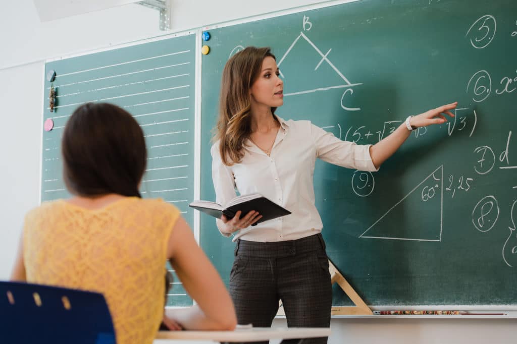 Teacher instructing at a chalkboard at the front of a classroom.