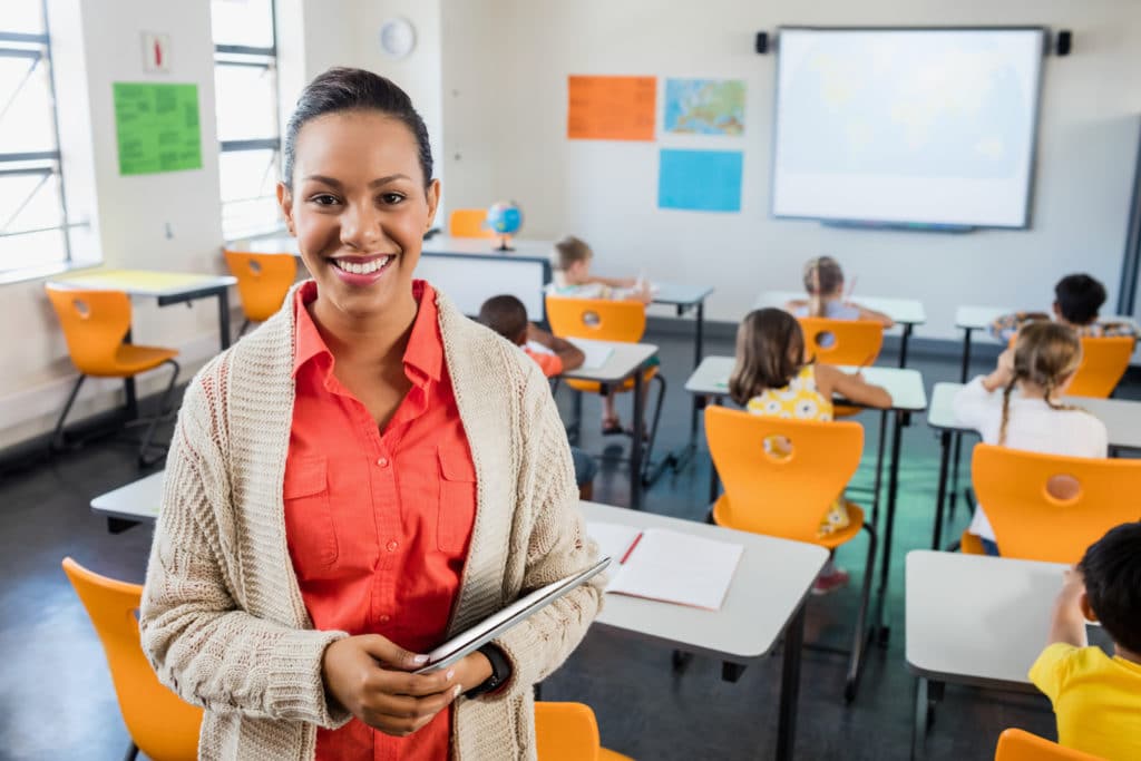 Smiling teacher standing at the back of a classroom holding a tablet.