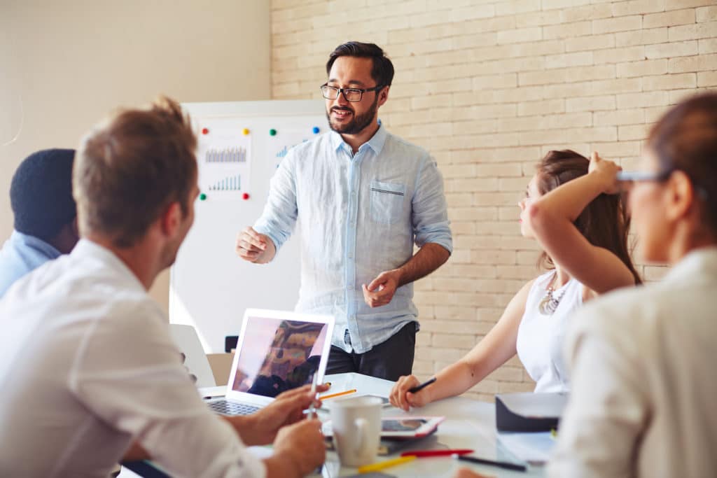Man standing by a table of colleagues explaining his idea.