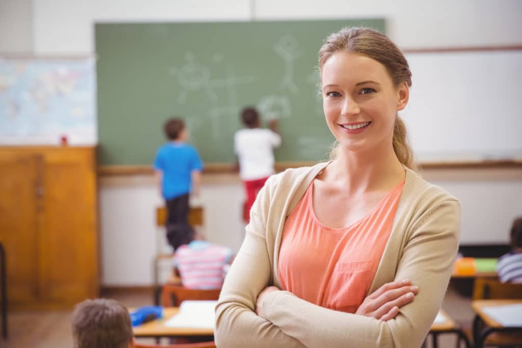 Young female teacher standing at the back of a classroom smiling.