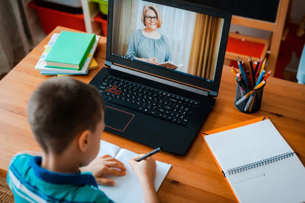Young writing on paper while listening to teacher instructing from laptop