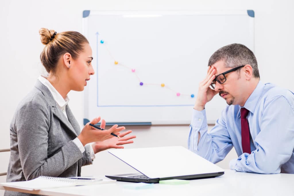 Woman talking to a bewildered man at a table with a laptop and notebook.