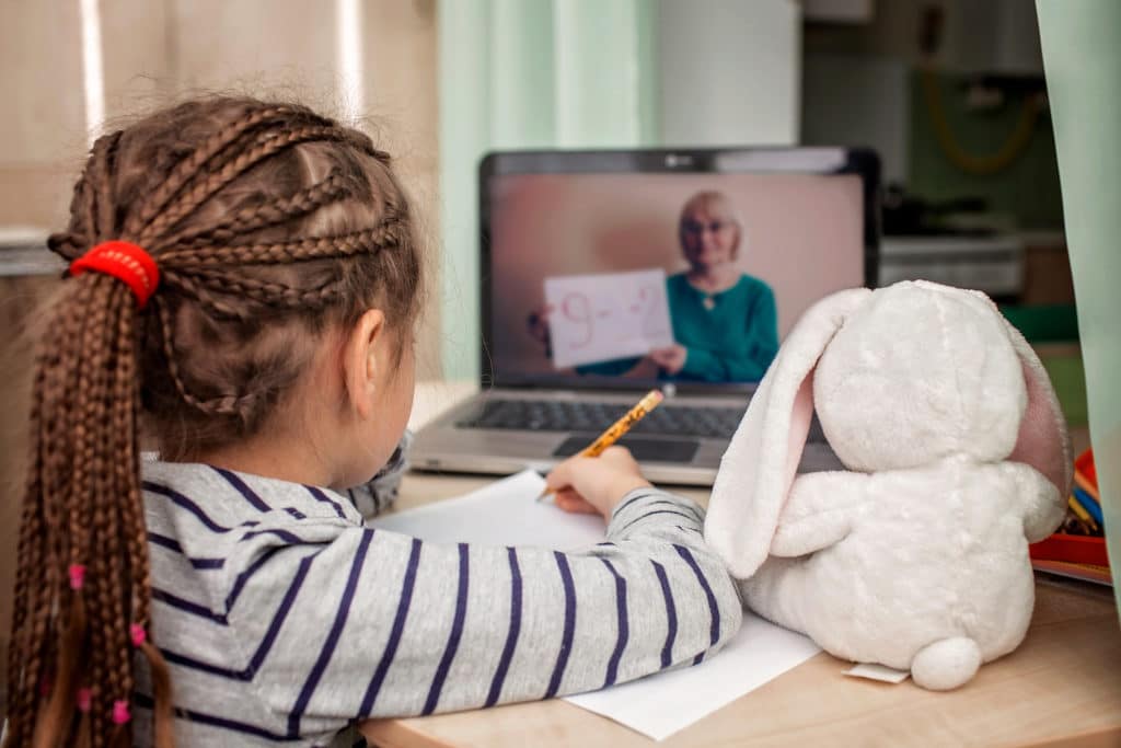 Young girl writing on a piece of paper while watching her teacher on a laptop.