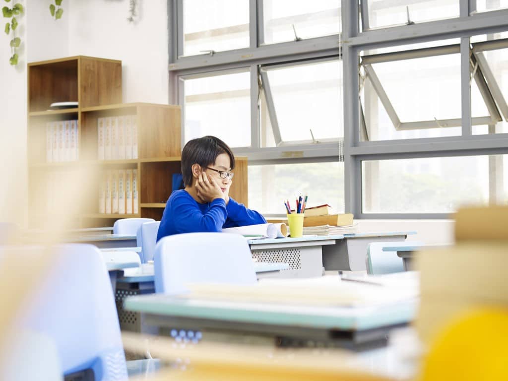 Young boy holding his head in his hands while sitting alone in a classroom.