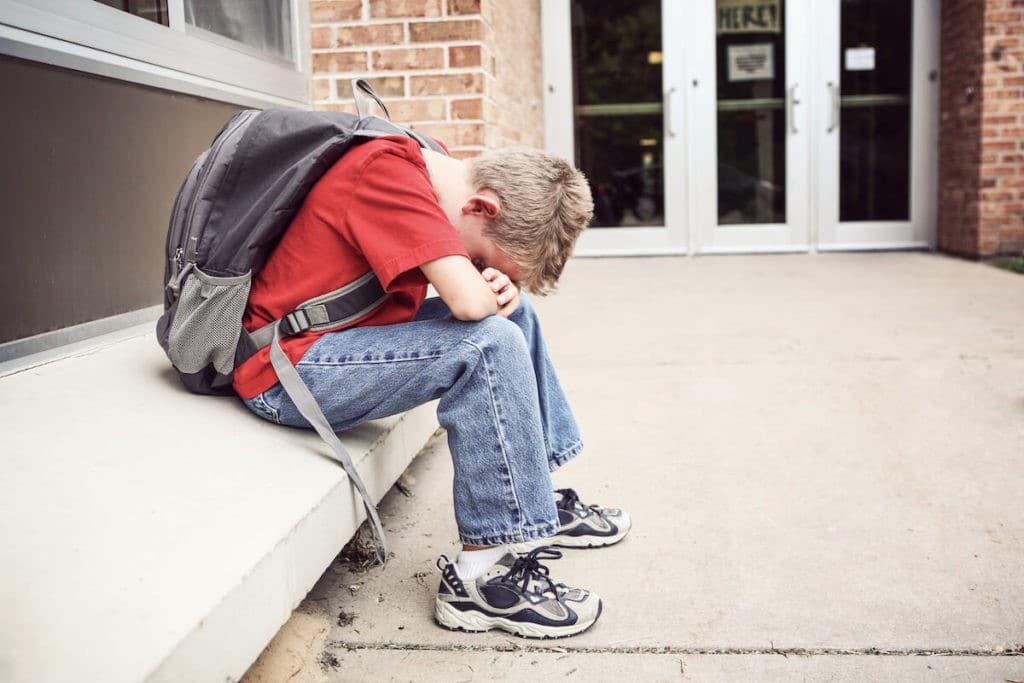 Young boy wearing a backpack with his head down on a school bench.