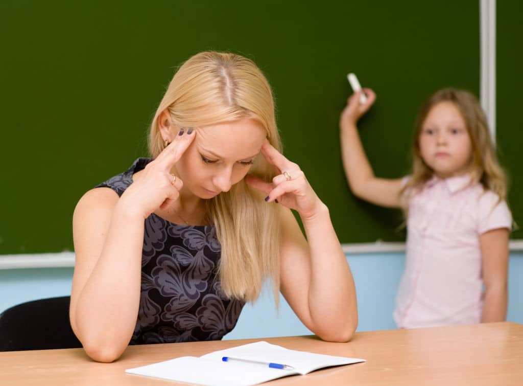 Teacher holding her head while sitting at her desk in a classroom.