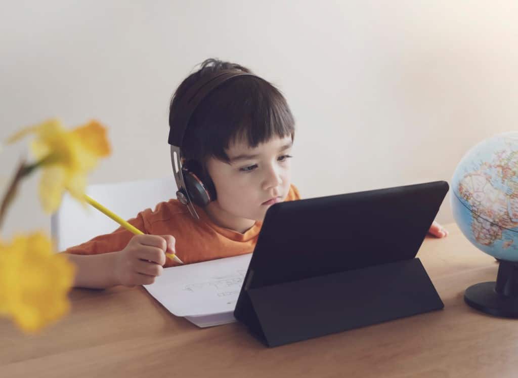 Young boy wearing headphones and writing while looking at a tablet.