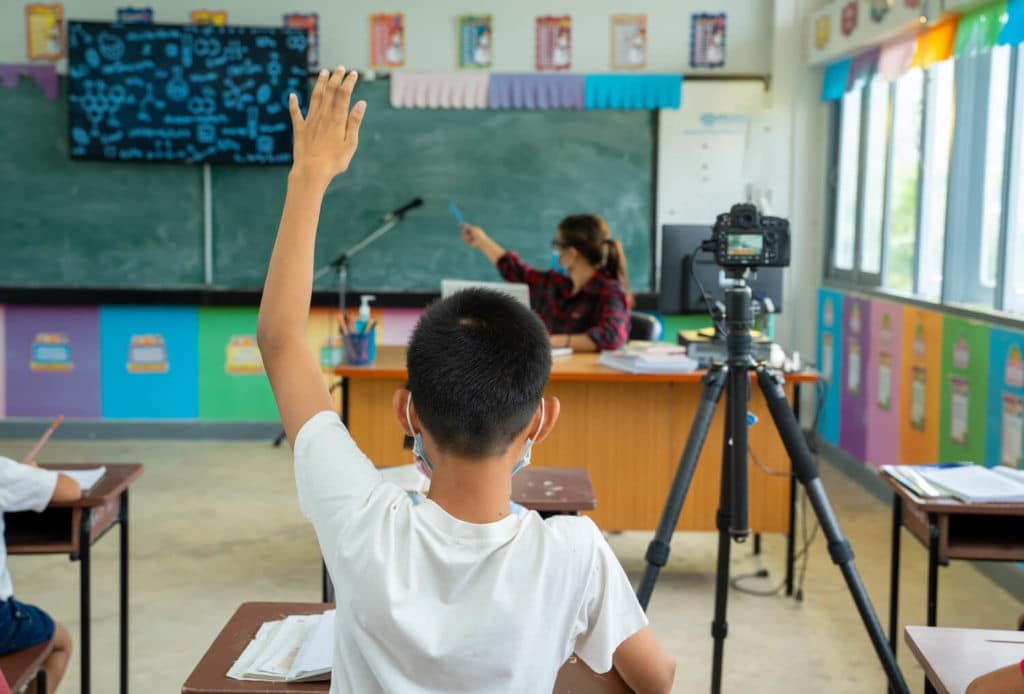 Classroom with young students and a teacher wearing masks and recording the lesson.