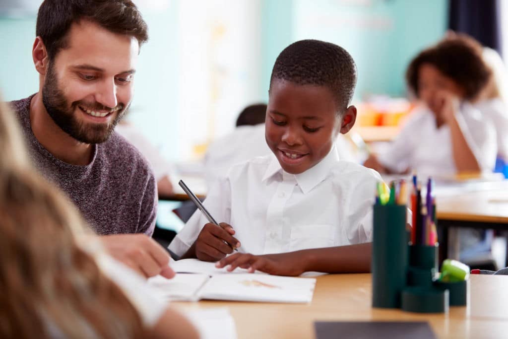 Teacher helping a young boy at his desk writing in a notebook.