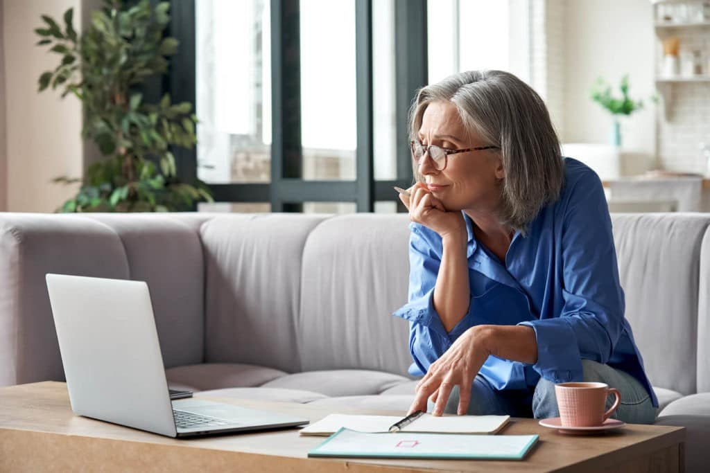 Older lady pondering while sitting on a couch and looking at papers and a laptop.