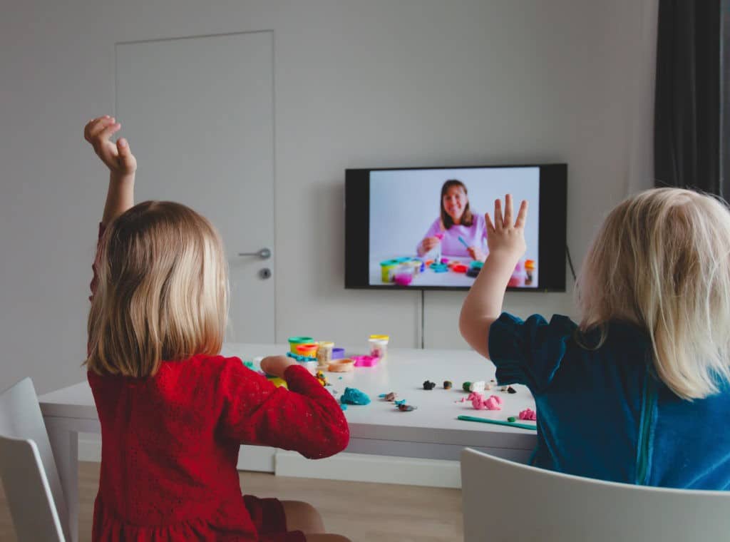 Two young girls sitting at a table playing with clay while watching a TV.
