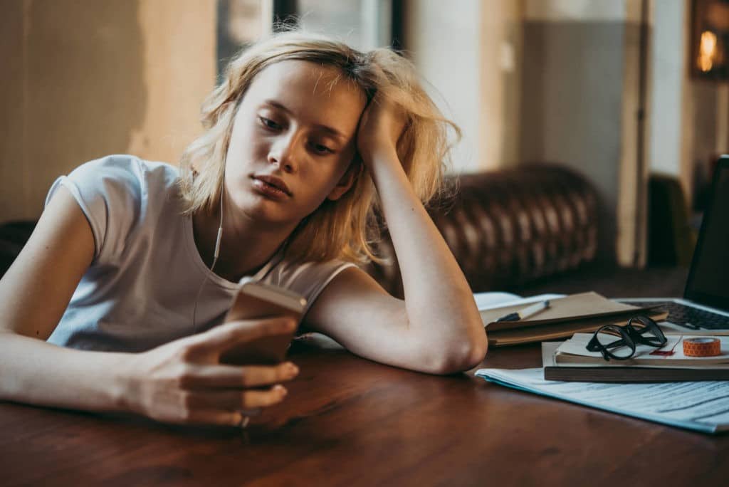 Bored girl on her smartphone while her work sits on the table.