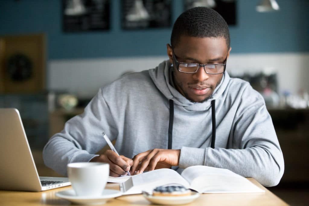 Young man sitting at a table taking notes with a book and laptop on the table.