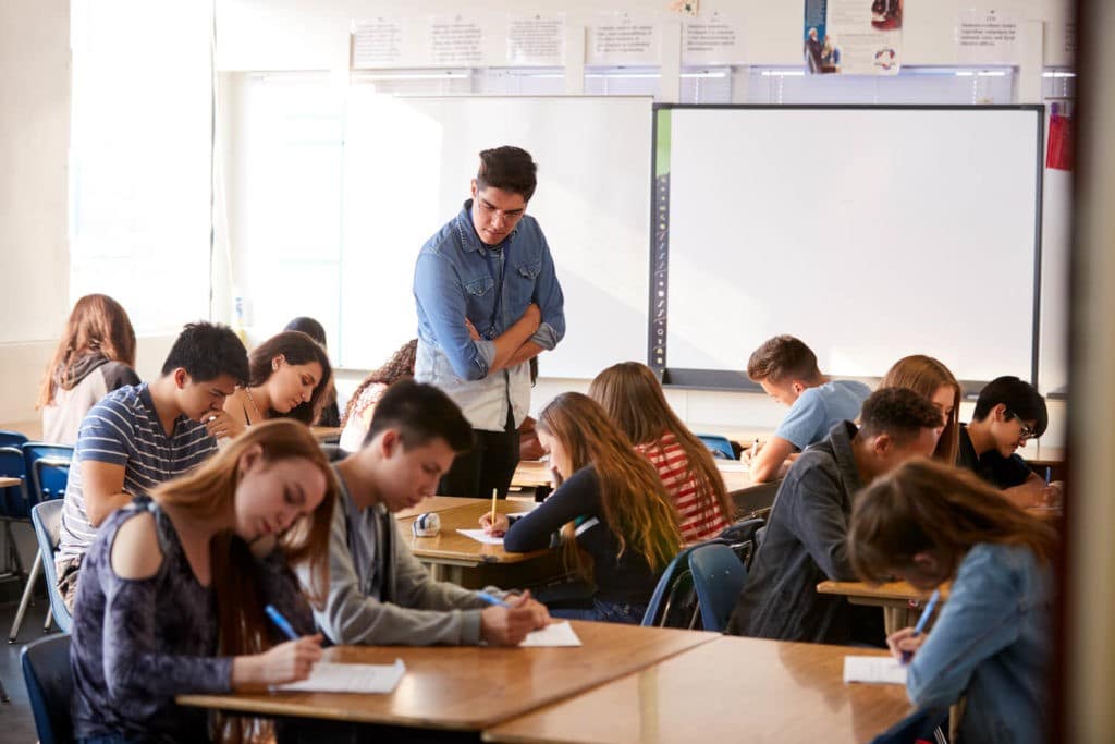 High school students working at their desks while a teacher walks and observes.