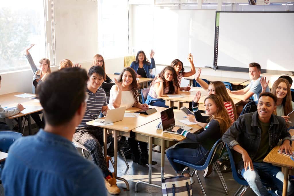 Classroom full of high school students with raised hands.