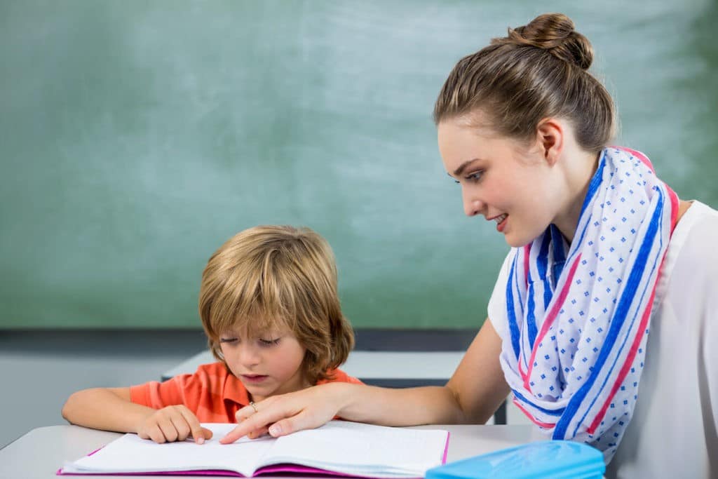 A teacher sitting at a desk with a young student helping him read.