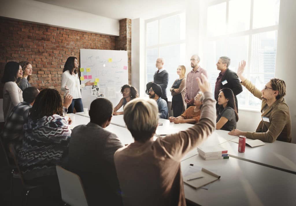 Employees in a group meeting raise their hands to ask questions.