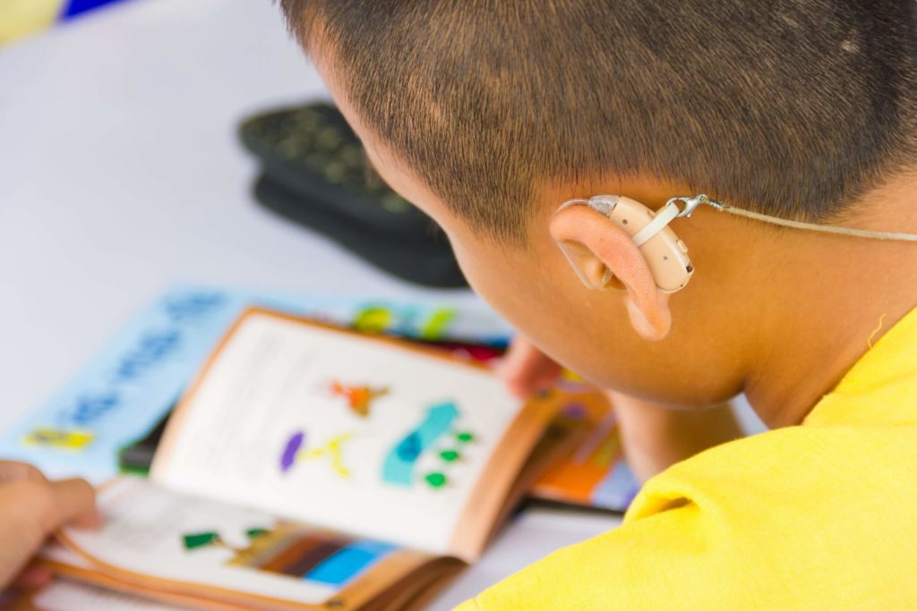 A close-up shot of a young boy wearing a hearing aid while reading a book in a classroom.