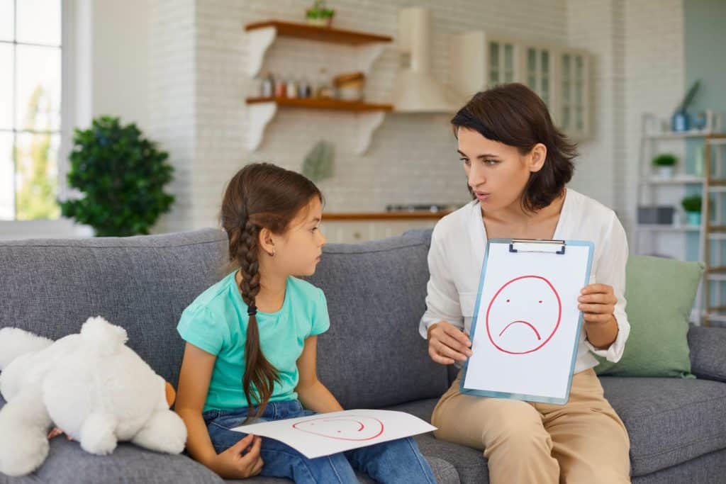 A child sits with a woman who is showing her a clipboard and a drawing of a frowning face.