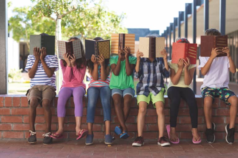 Front view of students reading behind books while sitting in a corridor.