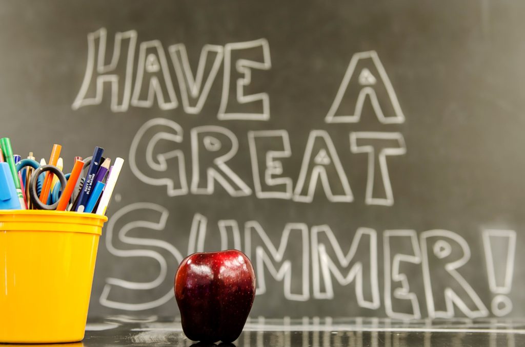 An apple and pencil holder sit on a teacher’s desk with “Have a great summer!” written on the chalkboard.