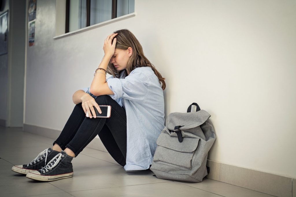 An upset teenage girl sits on the classroom floor, holding her head.