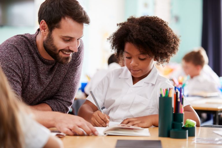 A male teacher works individually with a student at her desk.