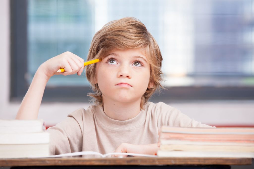 An elementary aged student sits at his desk and thinks, holding his pencil to his head.