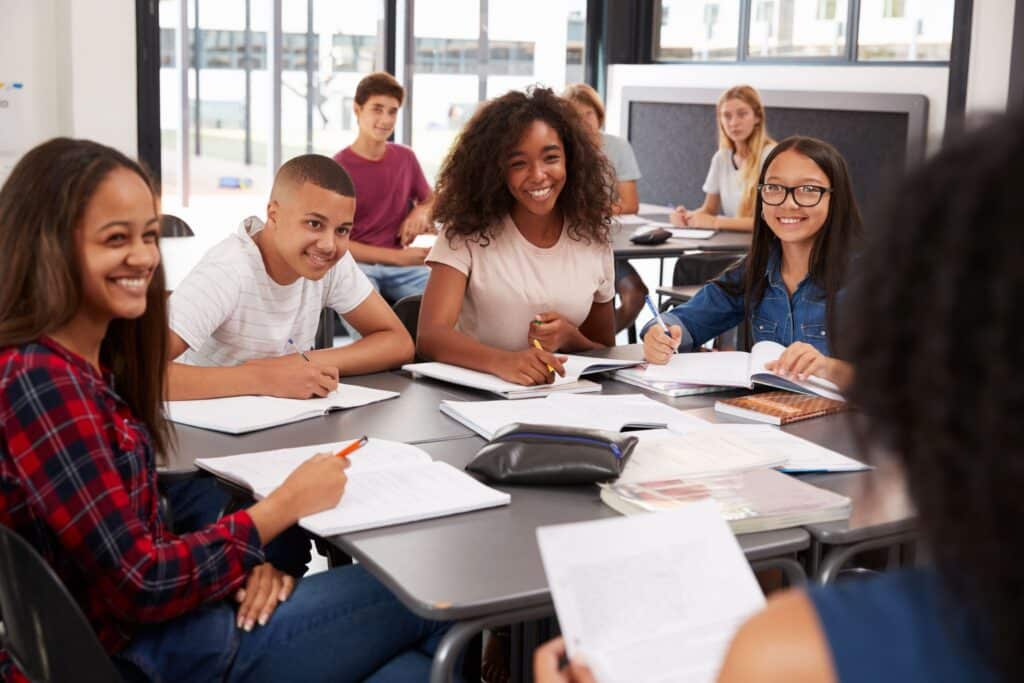 A group of high school students sit with their teacher at a table, discussing school work.