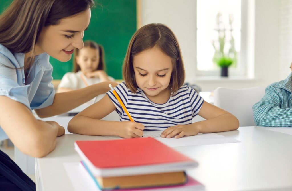 A teacher sits by a young student, helping her on an assignment.