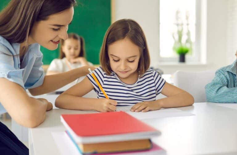 A teacher sits by a young student, helping her on an assignment.