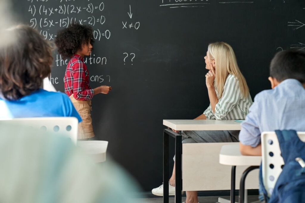 A student stands at the white board and talks with her teacher in middle grades mathematics class.