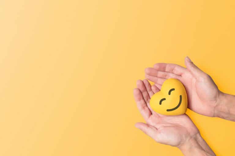 A pair of hands holds a yellow heart shaped stress ball that has a smiling face.