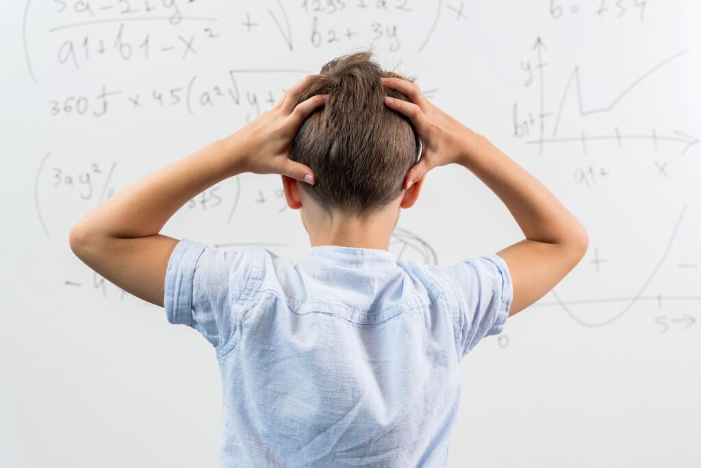 A student stands in front of the whiteboard with his hands on his head, thinking and problem-solving.