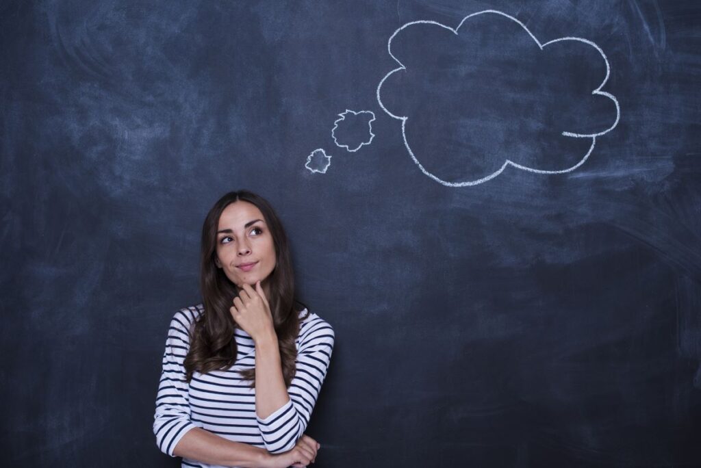 A woman stands in front of a chalkboard, thinking with a thought bubble written in chalk behind her.